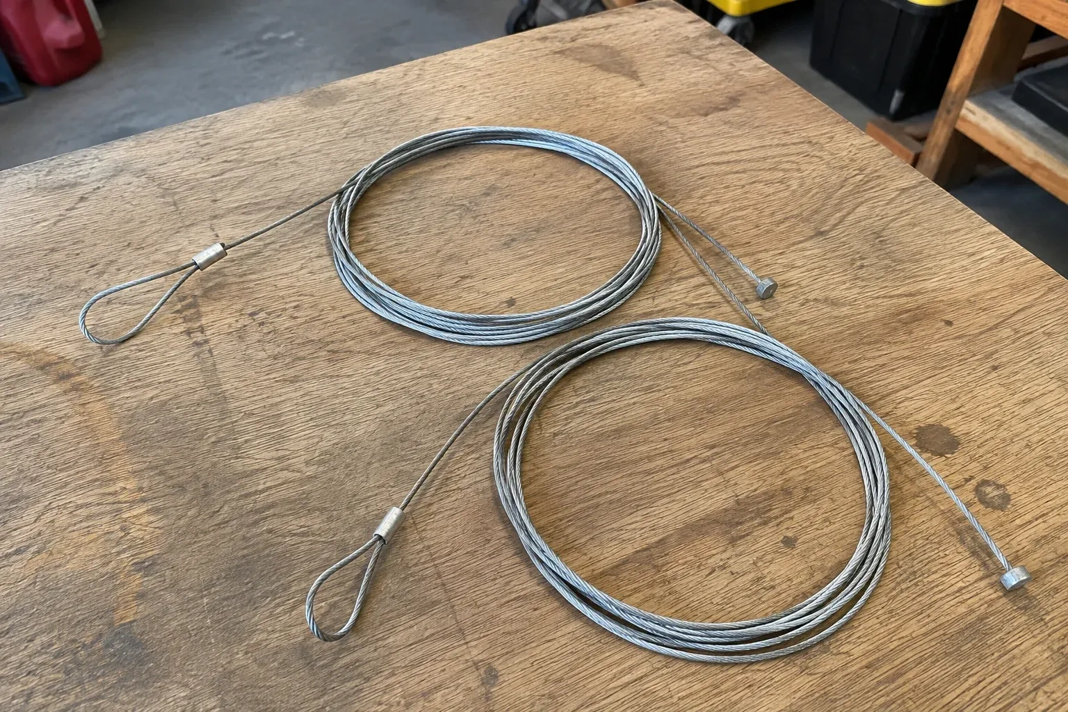 Two coiled steel cables with loop and stop ends on a workbench. — Garage Door Cable Replacement, Scugog