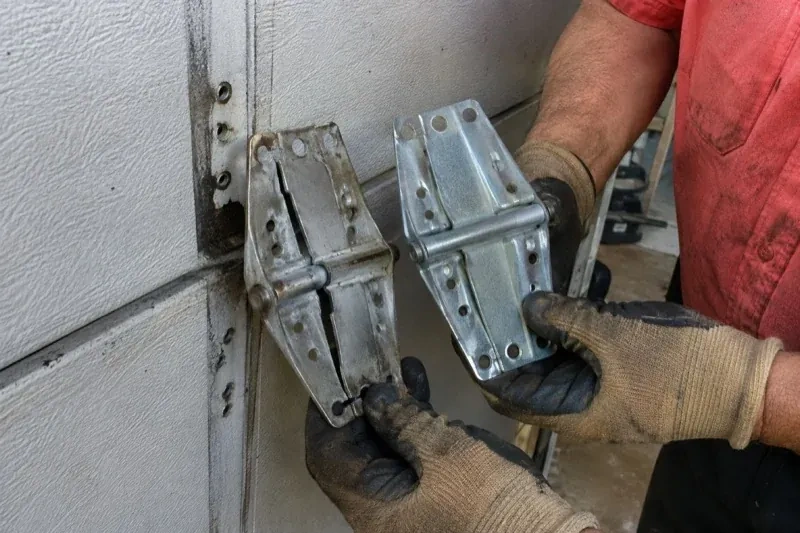 Gloved hands holding two metal garage-door hinges side by side, one discolored with a visible crack and the other bright silver. — Garage Door Hinge Replacement, Scugog
