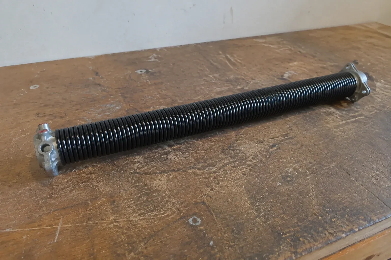 Detached black coil spring with metal end fittings on a workbench. — Torsion Spring Replacement, Scugog